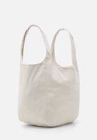 Torebka z miękkiego materiału w kolorze kremowym, z dwoma szerokimi rączkami, niestrukturalna i pojemna, stojąca na tle czystej białej ściany.