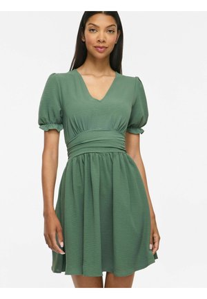 Femme portant une courte robe verte avec des manches bouffantes et une taille froncée, debout devant un fond blanc uni.
