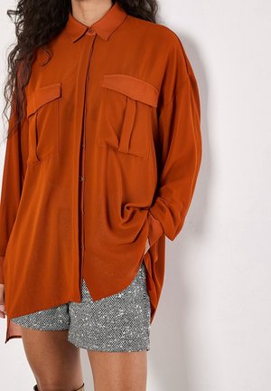 Kvinde iført en løstsiddende, brændt orange skjorte med store brystlommer og sort-hvide teksturerede shorts.