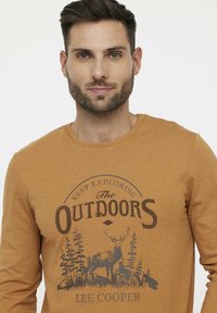 Langarm, mosterdkleurig T-shirt van zacht katoen met een buitenscène van bomen en een hert, gemarkeerd met "Lee Cooper."