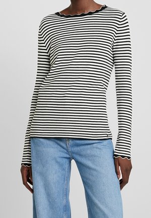 Personne portant un haut à manches longues à rayures horizontales noires et blanches avec des bords festonnés et un jean denim bleu clair.
