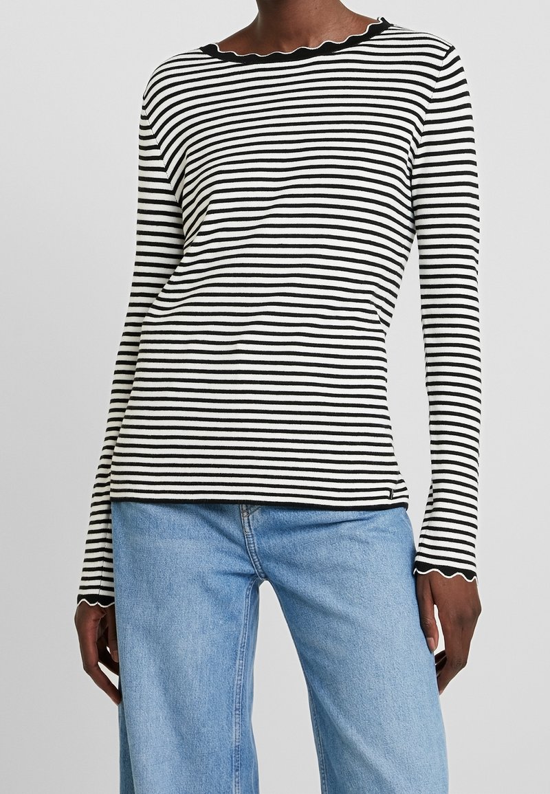Personne portant un haut à manches longues à rayures horizontales noires et blanches avec des bords festonnés et un jean denim bleu clair.
