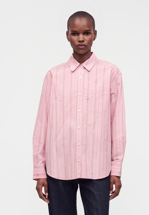 Personne aux cheveux courts portant une chemise rose ample à boutons avec des rayures verticales et un pantalon sombre, debout devant un fond uni.