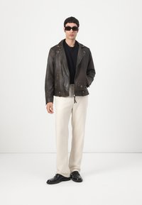Jachetă de motocicletă din piele maro cu fermoare argintii, tricou negru, pantaloni crem și pantofi negri, asortată cu ochelari de soare negri dreptunghiulari.