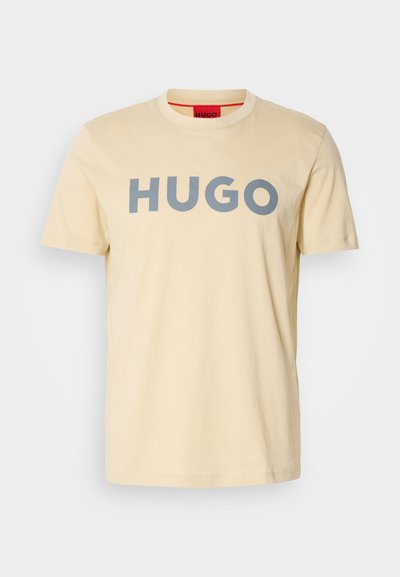 Светло бежова памучна тениска с къси ръкави, с голям сив логотип "HUGO" отпред и червен акцент на вътрешната яка.