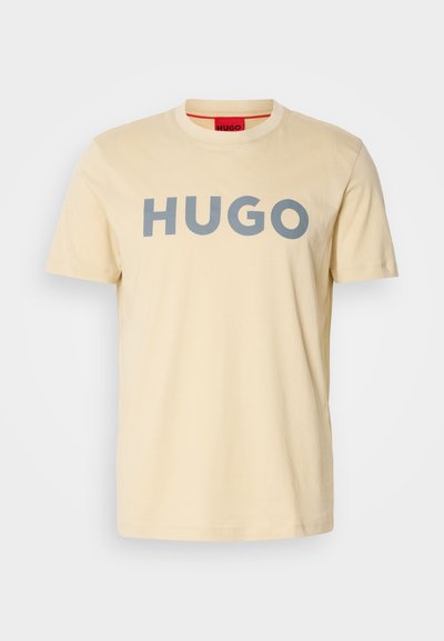 Светло бежова памучна тениска с къси ръкави, с голям сив логотип "HUGO" отпред и червен акцент на вътрешната яка.