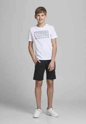 Camiseta blanca con estampado de logo gris, pantalones cortos negros con bolsillos y zapatillas blancas. El modelo posa con una mano en el bolsillo, sonriendo.