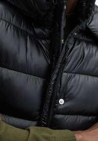 Czarna kurtka puchowa z zamkiem z przodu, pikowana faktura i wysoki kołnierz. Srebrny zatrzask na piersi, lekka i ocieplana.