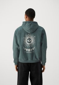Sudadera verde con un diseño gráfico que presenta un ojo, patrones celestiales y texto. Tela suave con un corte relajado y capucha con cordón.