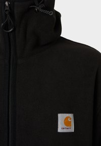 Must mustaga, millel on kõrge krae, ees tõmblukk ja seotav strings. Vasakul rinnal on ruudukujuline silt, millel on oranž logo.