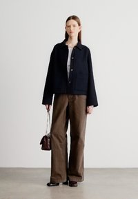 Casaco azul-marinho curto com mangas largas, bolsos frontais e fecho de botões, combinado com calças castanhas de perna larga e uma pequena mala escura.