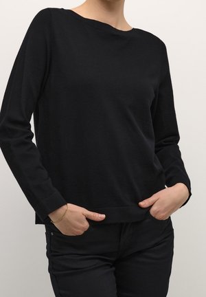 Osoba ubrana w prostą czarną bluzkę z długim rękawem i czarne spodnie, z rękami włożonymi do przednich kieszeni, z złotą bransoletką na lewej ręce.