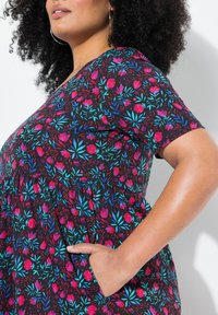 Kukkamekko, jossa on musta tausta ja elävät vaaleanpunaiset ja siniset kukkakuviot, lyhyet hihat ja sivutaskut. Tekstuuroitu kangas.