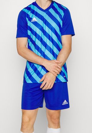Homme portant un uniforme de football Adidas bleu avec des rayures diagonales bleu clair sur le maillot et un short bleu uni, debout les mains jointes.