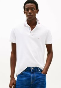 Jonge man in een witte korte mouwen polo met een klein logo en een blauwe spijkerbroek, hand in de zak, neutrale uitdrukking, witte achtergrond.