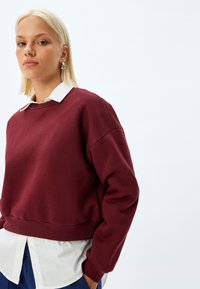 Bordowy sweter z ribbowanym dołem i manszetami, noszony na białej koszuli z kołnierzykiem. Gładka faktura i lekko skrócony fason.