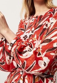 Blouse à motif floral rouge avec de longues manches ballons, présentant un design à boutons et des poignets élastiqués. Fabriquée en tissu léger et texturé.