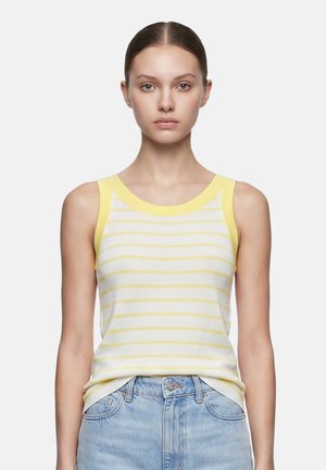Giovane donna dalla pelle chiara e capelli tirati indietro che indossa un top senza maniche a righe gialle e bianche e jeans azzurri su sfondo bianco.