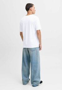 Mann, der von der Kamera weg blickt, trägt ein schlichtes weißes T-Shirt mit kurzen Ärmeln, lockere hellblaue Jeans und schwarze Slippers vor einem weißen Hintergrund.