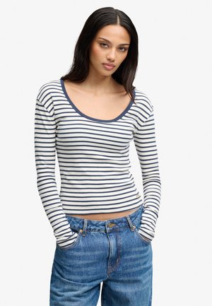 Mujer con cabello largo y oscuro, vestida con una camiseta de manga larga blanca con rayas azul marino y vaqueros azules, con las manos en los bolsillos, sobre un fondo liso.