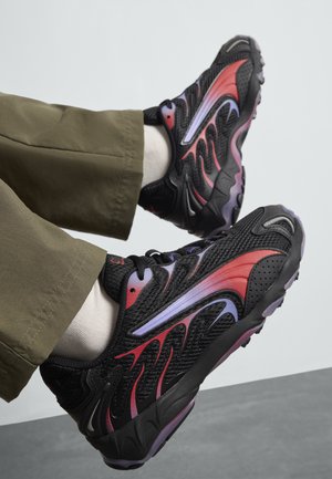 Black athletic shoes with flame-like red and purple accents, textured mesh upper, lace closure, and rubber sole. Worn with olive pants.