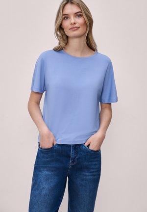 Frau trägt ein hellblaues Kurzarmshirt und blaue Jeans, die Hände in den Taschen, steht vor einem einfarbigen beigen Hintergrund.