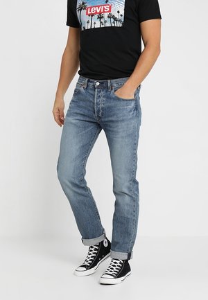 Mężczyzna w jasnoniebieskich podwiniętych dżinsach, czarnych wysokich trampkach i czarnej koszulce Levi's z grafiką palmy.