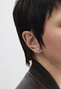 Silver heart-shaped earring with a faceted pink gemstone in the center. Close-fitting design, worn on the left ear against short dark hair.