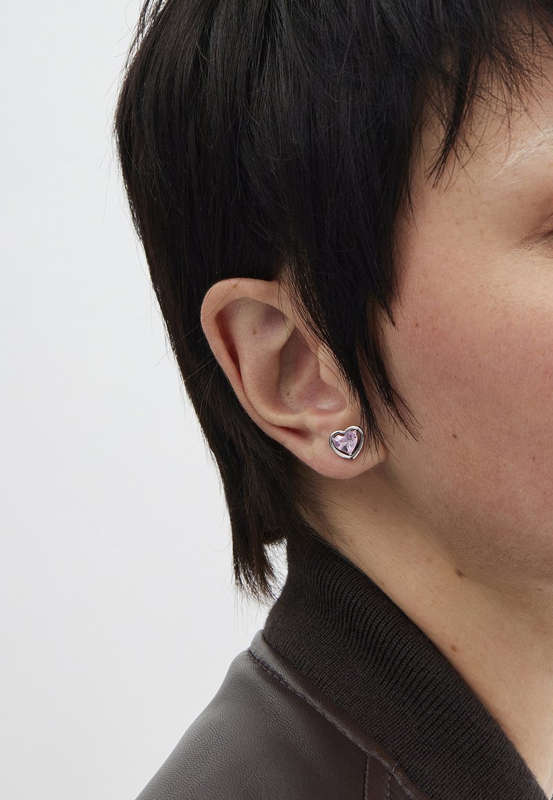 Silver heart-shaped earring with a faceted pink gemstone in the center. Close-fitting design, worn on the left ear against short dark hair.