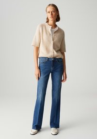 Lichtbeige gebreide cardigan met korte mouwen en knopen, gecombineerd met blauwe uitlopende jeans. Witte schoenenaccenten maken de look af.