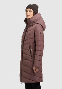 Pufferjakke i mauve med skrå quiltet mønster, lynlås foran, justerbar hætte og sidelommer. Modellen bærer en mørk beanie.