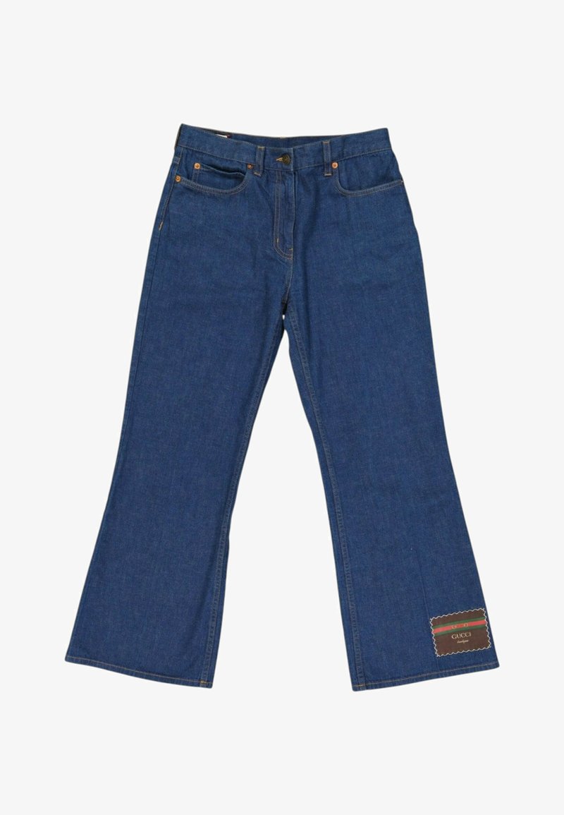 Jean large en denim bleu avec poches avant et arrière, fermeture par boutons, et un patch étiquette Gucci près de l'ourlet inférieur sur une jambe.