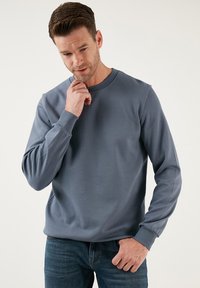 Felpa blu chiaro in misto cotone, collo rotondo, tessuto a textura, polsini a costina, abbinata a jeans scuri, su sfondo neutro.