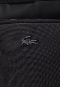 Tissu texturé noir avec un petit logo crocodile noir embossé sur le devant, au-dessus d'une couture cousue.