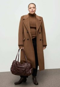 Bruine wollen mantelmix met zwarte knopen, geribbelde bruine trui, zwarte broek, bruine leren handtas met ritsen en enkellaarsjes.