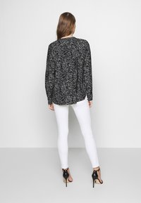 Blouse noire à manches longues avec un motif abstrait blanc, associée à un jean blanc ajusté et des sandales à talons hauts noires. Vue de dos.