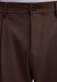 Bruine op maat gemaakte broek van een gladde stof, met een knoopsluiting, zijzak en scherpe naden. De stof is lichtgewicht en gestructureerd.