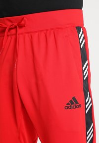 Pantalons de survêtement rouges Adidas avec des panneaux latéraux rayés noir et blanc, taille avec cordon de serrage, et logo sur la cuisse.