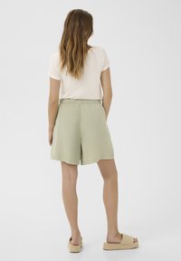 Šviesiai žali, iki kelių siekiantys šortai su elastine juosta ir šoninėmis kišenėmis, derinami su kreminiu trumpomis rankovėmis marškinėliais ir smėlio spalvos šlepetėmis.