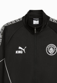 Czarna kurtka sportowa z wysokim kołnierzem, zamkiem błyskawicznym, białymi akcentami oraz bocznymi panelami ze wzorem geometrycznym. Na niej widnieje napis "KING" oraz logo Manchester City.