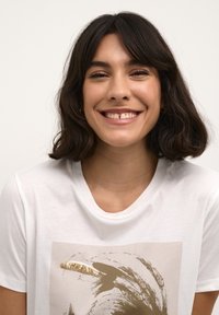 T-shirt blanc avec un motif beige orné de sequins dorés, encolure ronde et manches courtes, porté par une personne souriante aux cheveux foncés.