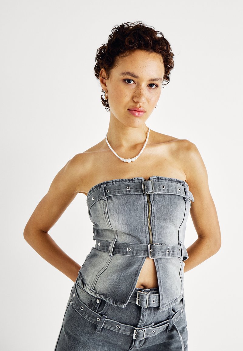 Top sin mangas de mezclilla gris con múltiples acentos de cinturón, un zipper frontal y detalle recortado. Tejido texturizado con silueta ajustada.