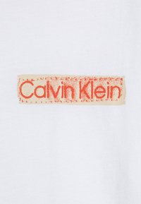 Camiseta de algodón blanca con una etiqueta rectangular beige con letras naranjas que dicen "Calvin Klein". Textura suave, diseño casual.