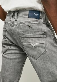 Jeans de mezclilla gris con un diseño clásico de cinco bolsillos, costuras prominentes, una etiqueta de marca azul en la cintura y tela texturizada.