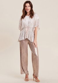 Cardigan di paillettes rosa chiaro scintillante con maniche larghe e vita con coulisse, abbinato a pantaloni beige plissettati a gamba larga iridescenti.