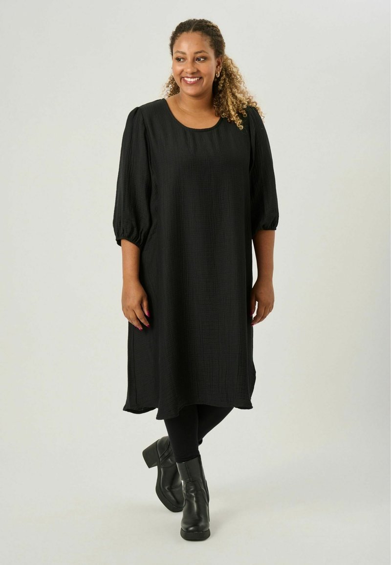 Vestido negro de tela texturizada con un corte holgado, escote redondo y mangas abullonadas. Se lleva con leggings negros y botas de tobillo.