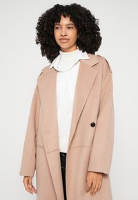 Beige ullrock med skräddarsydd design, slagkragar och en enda svart knapp, bärs över en vit skjorta och ljus polotröja.