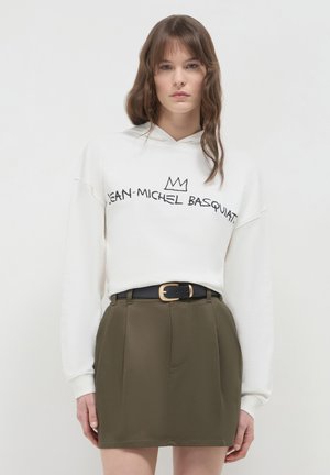 Femme portant un sweat à capuche blanc avec le texte "Jean-Michel Basquiat" et un graphique de couronne, associé à une jupe vert olive et une ceinture noire.