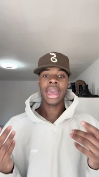 Young man wearing brown cap and light hoodie gestures with hands while speaking indoors, boots on shelf in background.
