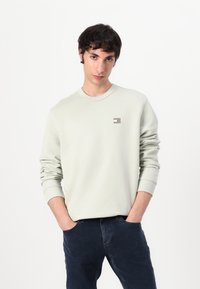 Felpa verde chiaro con collo a giro, polsini a costine e una piccola etichetta sul petto. Indossata con jeans blu scuro, mani nelle tasche.